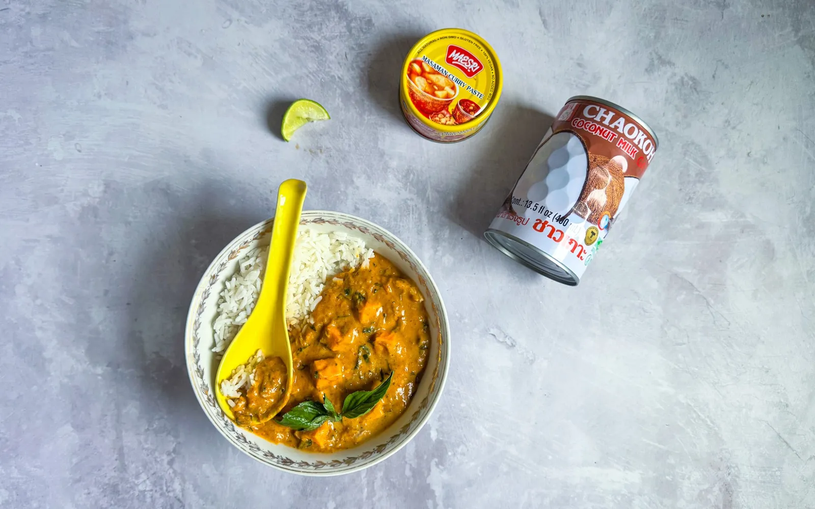A bowl of massaman curry with chicken, squash, and Thai basil over jasmine rice, next to a can of Mae Sri massaman curry paste, a can of Chaokoh coconut milk, and a lime wedge