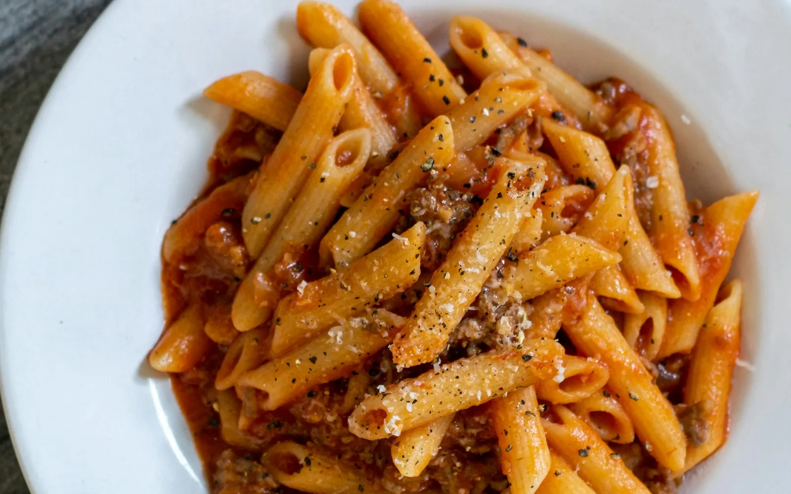 A white bowl of penne in a rich tomato meat sauce on a weathered wooden table
