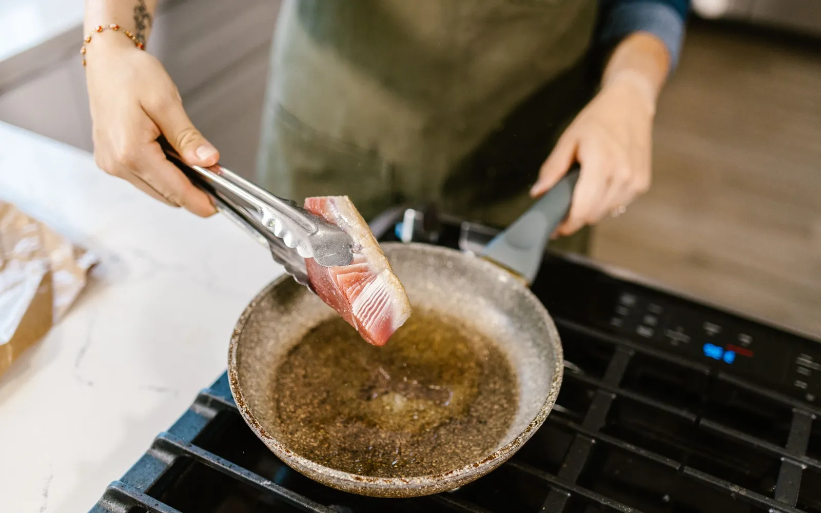Tongs lifting a seared tuna steak out of a skillet of hot oil on a gas stove
