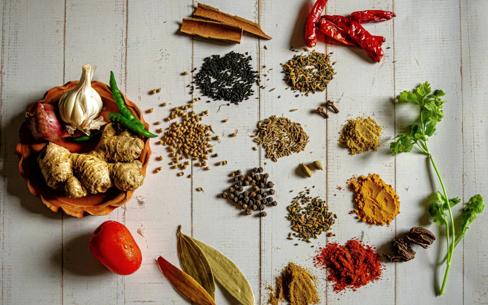 Whole and ground spices in small piles on a white wooden table, with garlic, ginger, chiles, bay leaves, and cilantro
