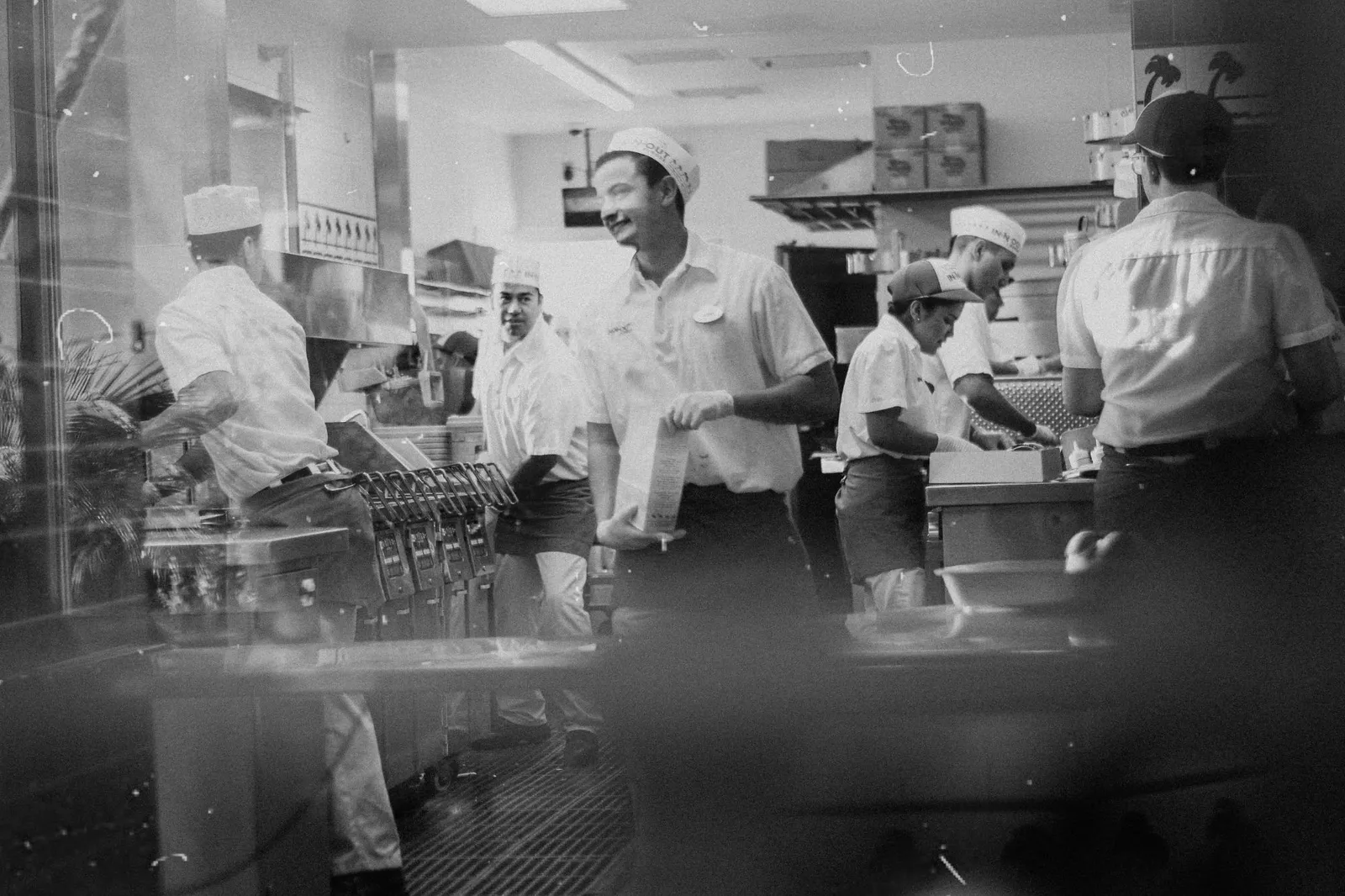 Black-and-white photo of a busy fast-food kitchen crew in paper hats, seen through the window